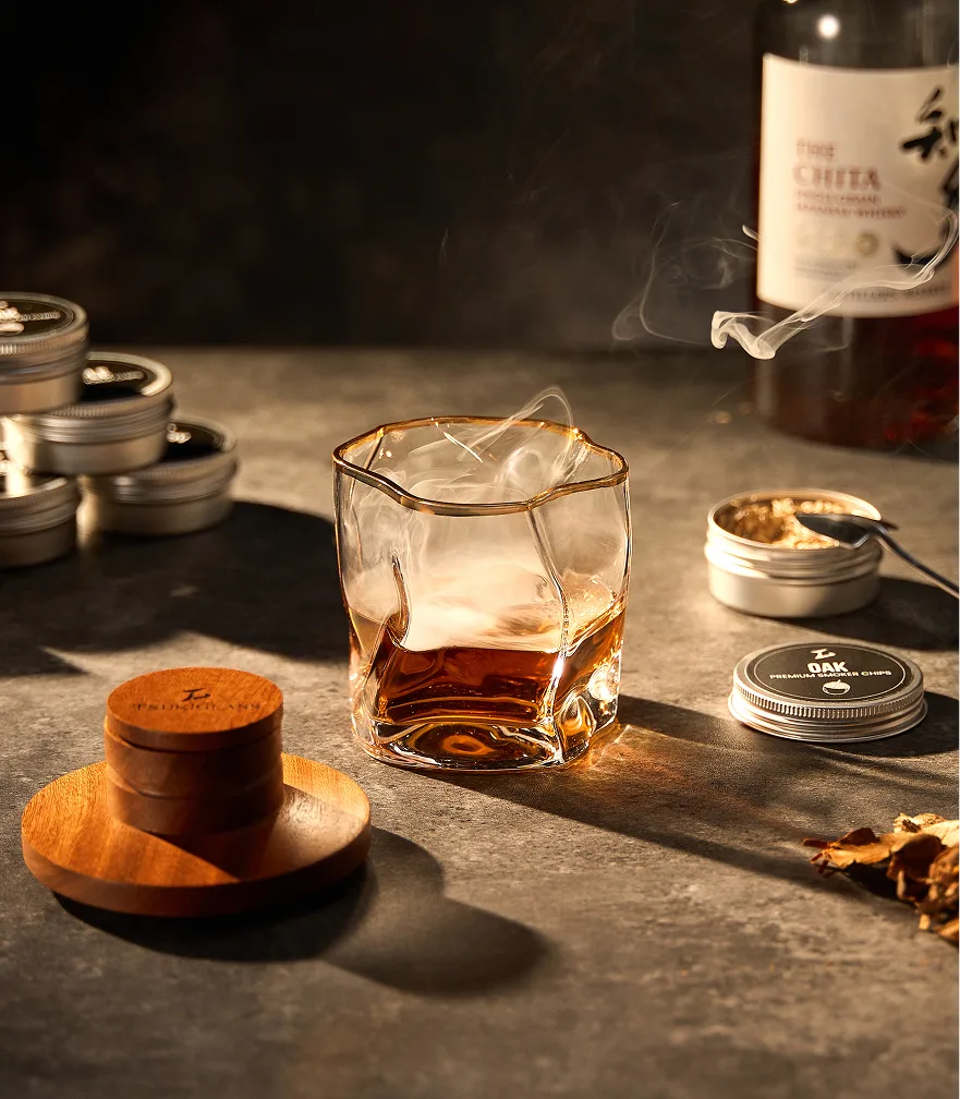 A glass of smoking whiskey sits on a table next to a bottle and tins of wood chips.