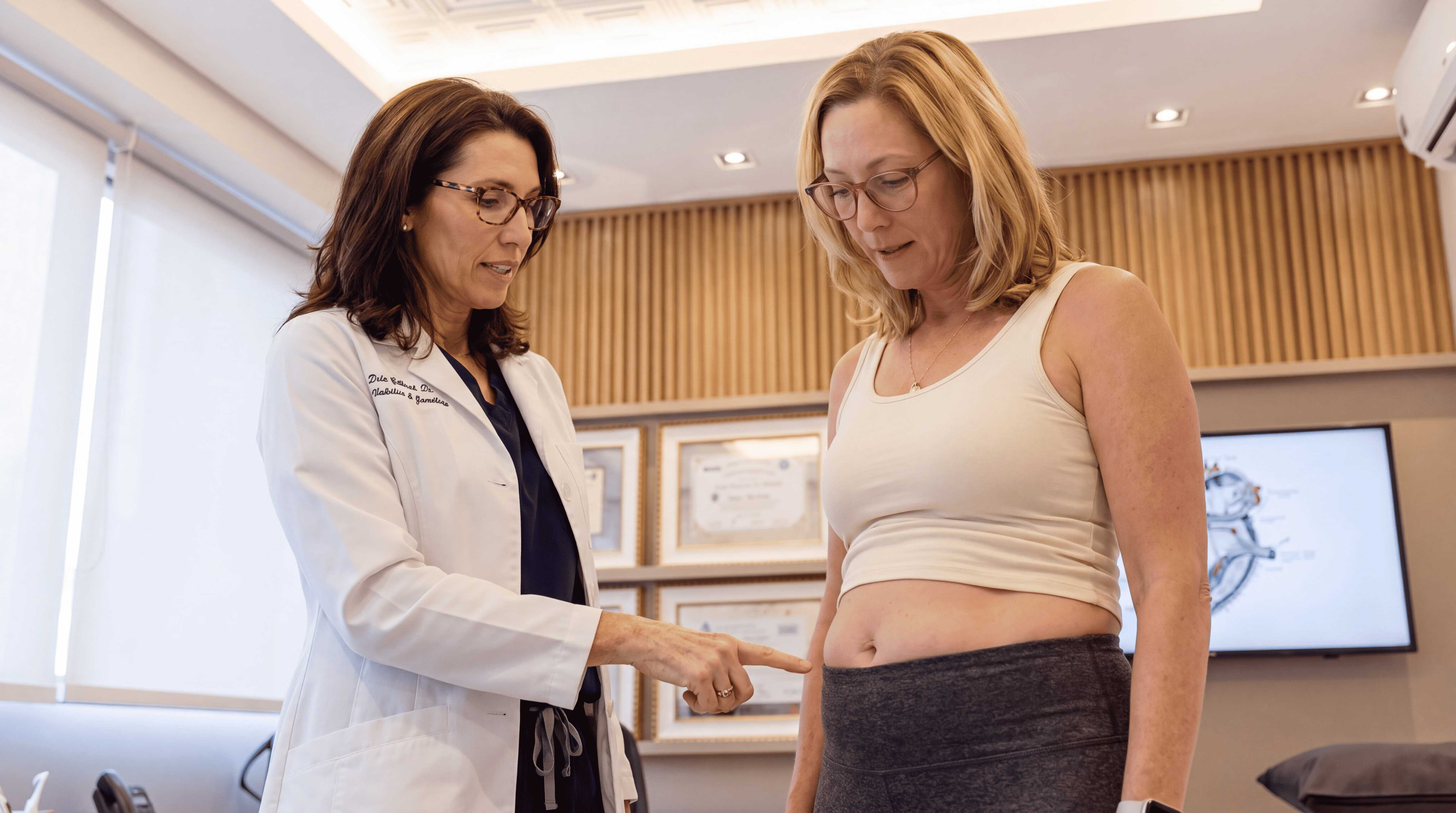 A female doctor in a white lab coat points to a patient's abdomen during an examination.