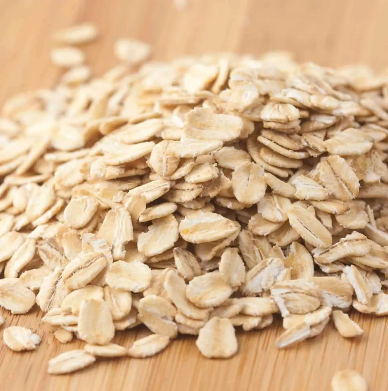 A close-up of a pile of uncooked rolled oats on a light brown wooden surface.