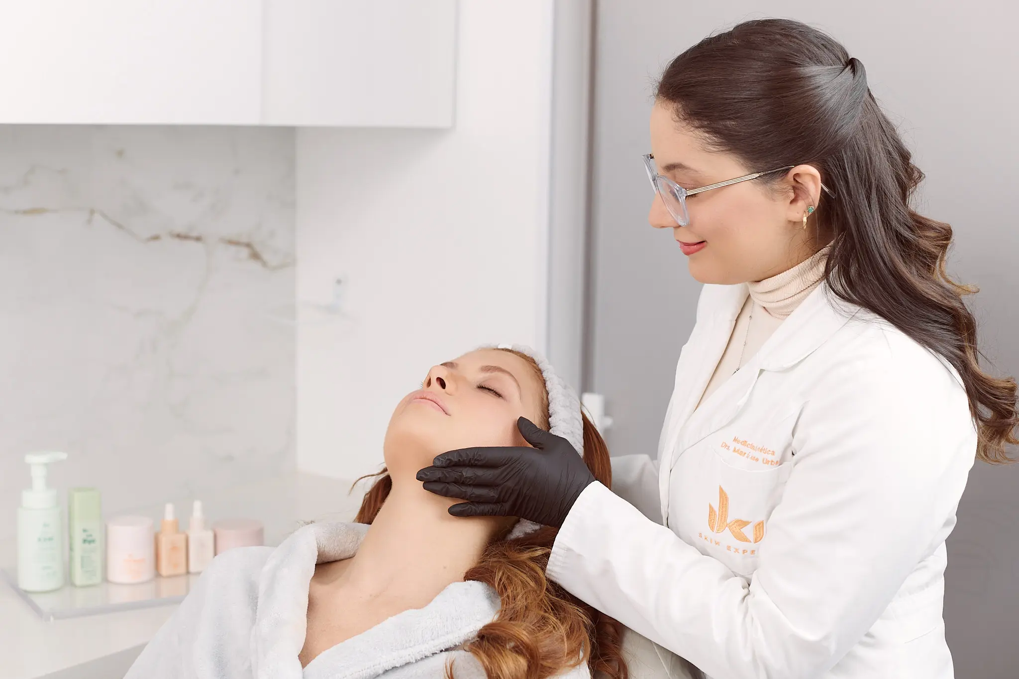 An aesthetician in a white coat and black gloves examines a relaxed woman's face during a skincare treatment.