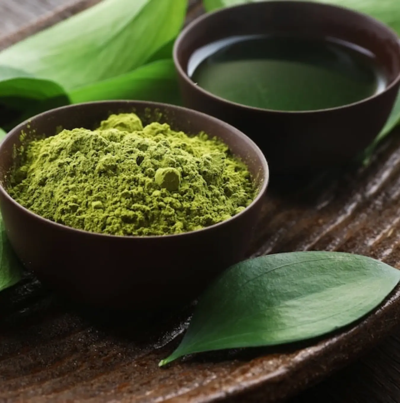 A bowl of green powder and a cup of green liquid on a wooden tray with fresh leaves.