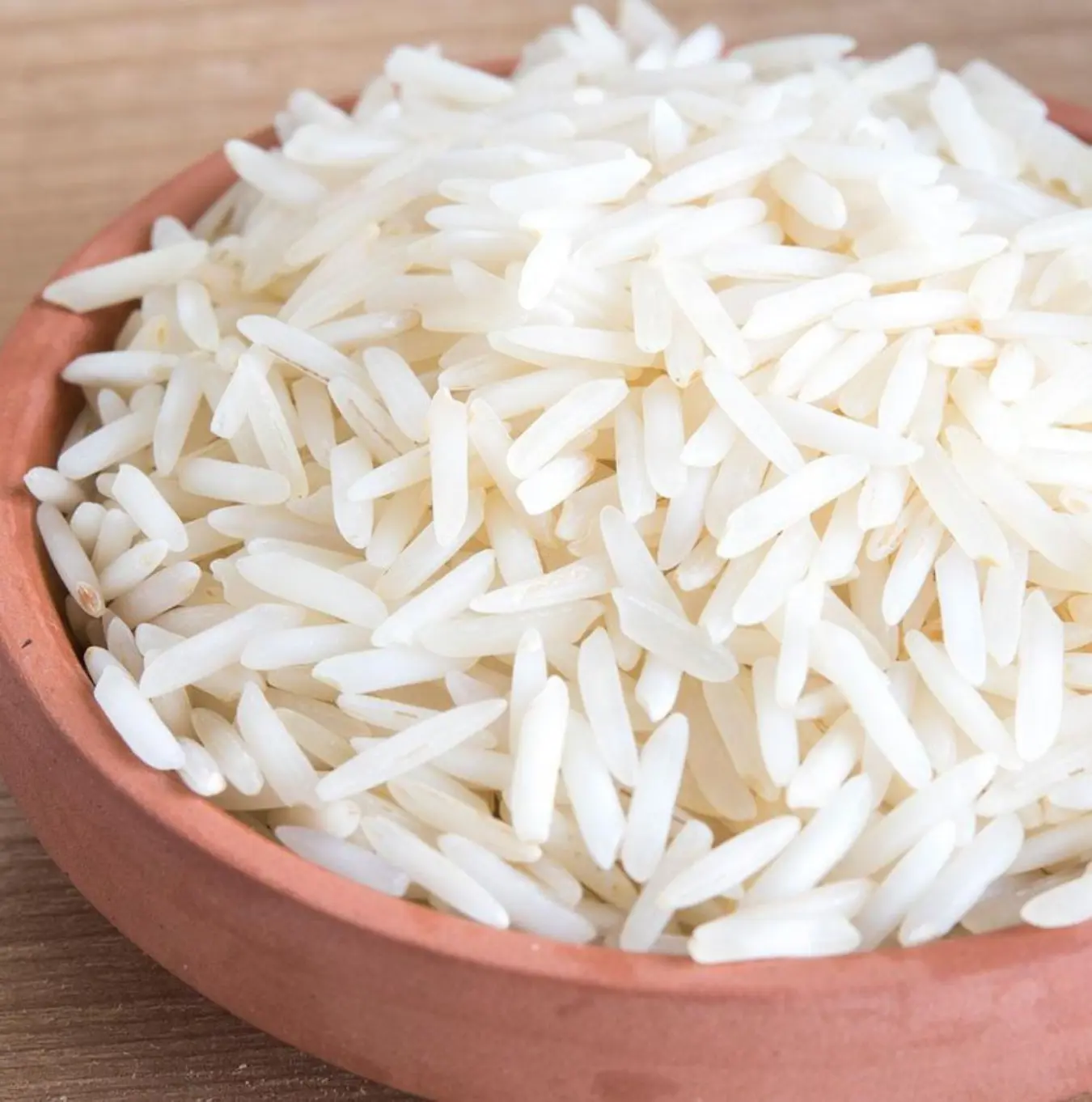 A close-up of uncooked long-grain white rice in a terracotta bowl on a wooden surface.