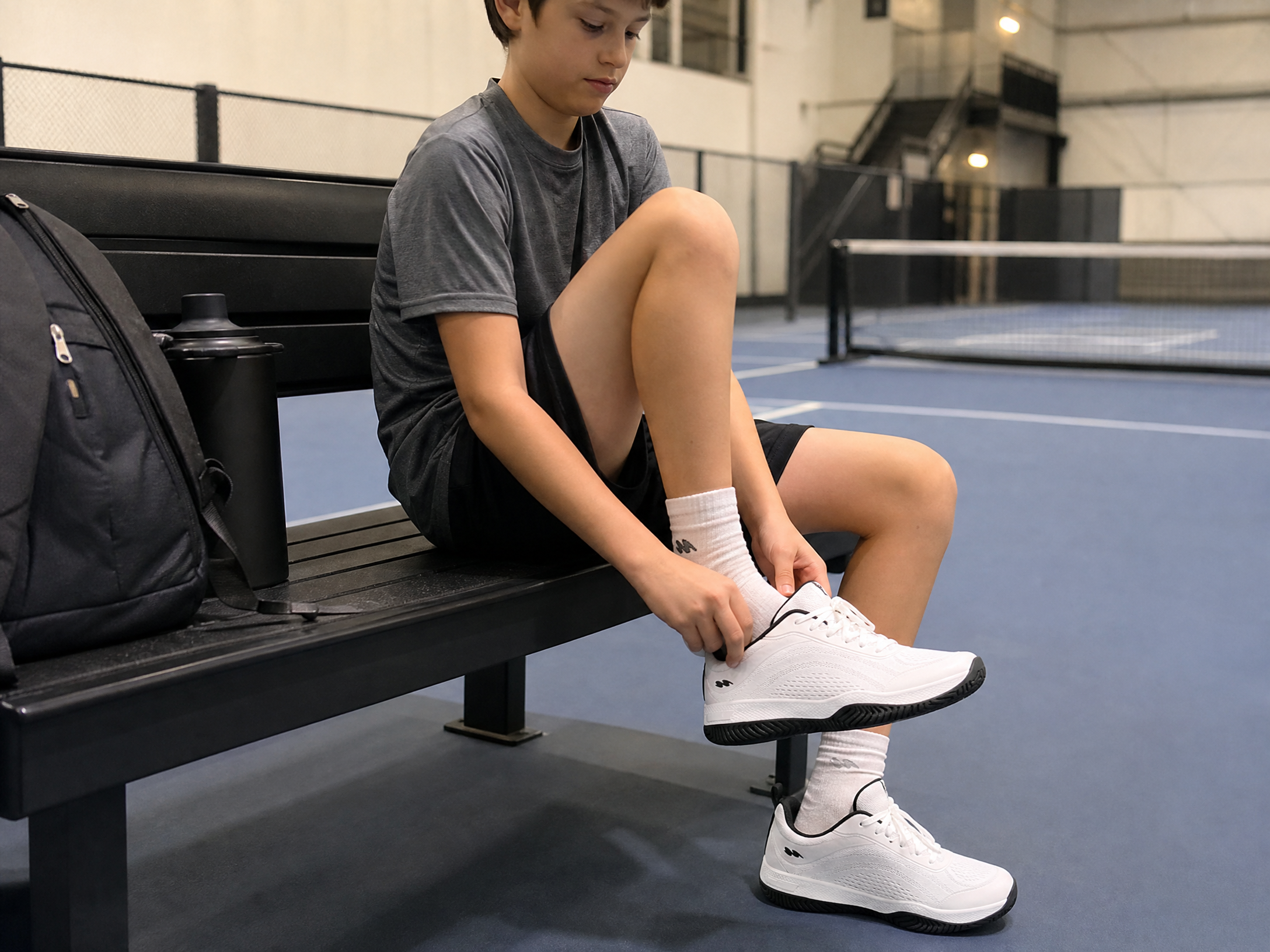 A boy sits on a bench on an indoor court, putting on his white athletic shoes.