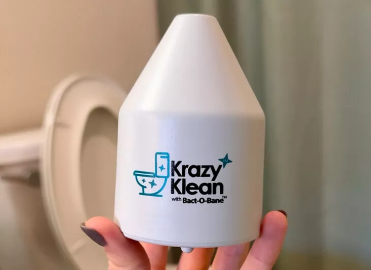 A hand holds a white Krazy Klean automatic toilet bowl cleaner in front of a toilet.