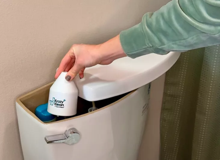 A person placing a Krazy Klean toilet cleaner into the tank of a toilet.