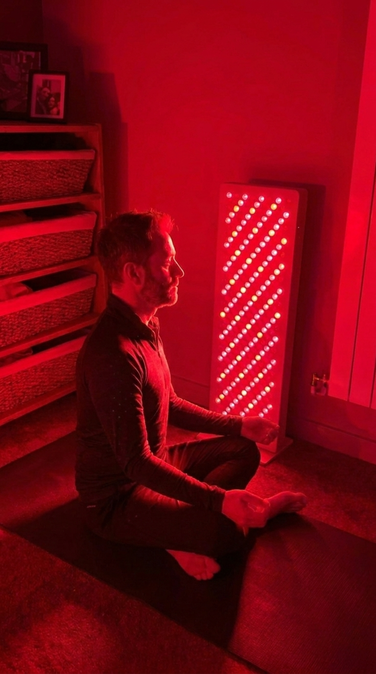 A man sits cross-legged on a yoga mat next to a tall red light therapy panel, meditating.