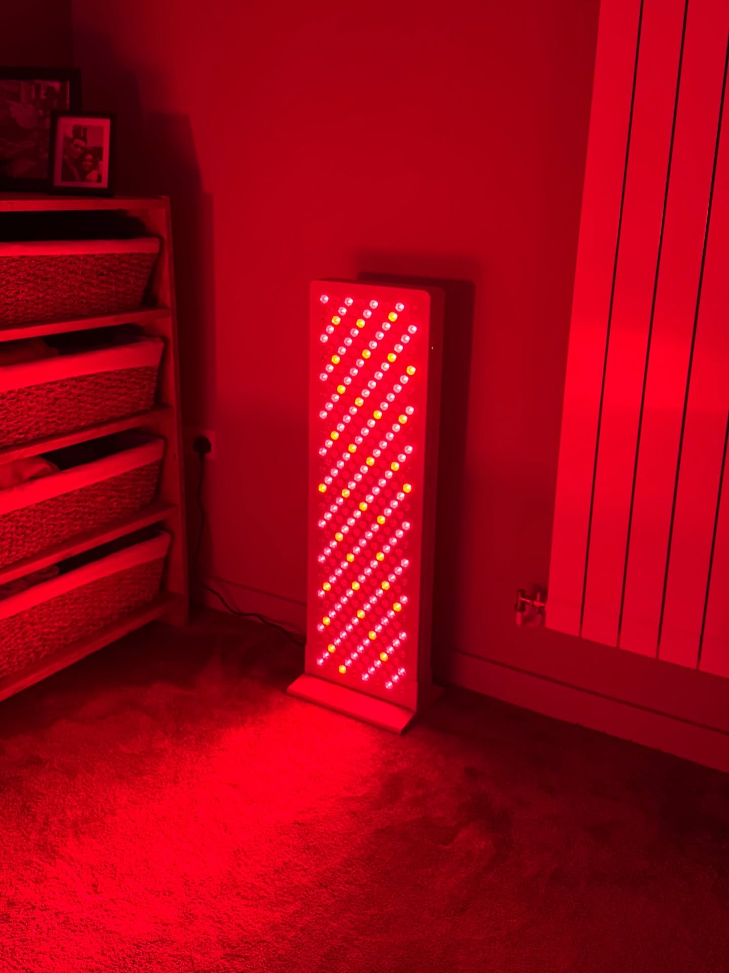 A red light therapy panel stands on the floor, casting a bright red glow in a room.