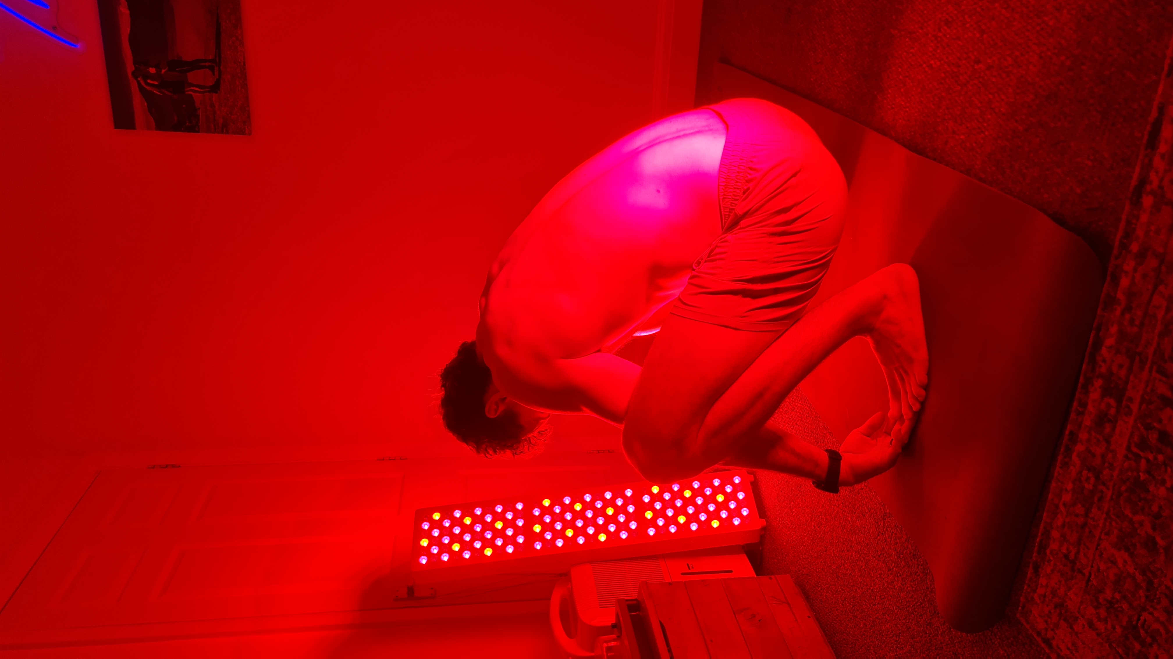 A person squats on a yoga mat in a room illuminated by a red light therapy panel.