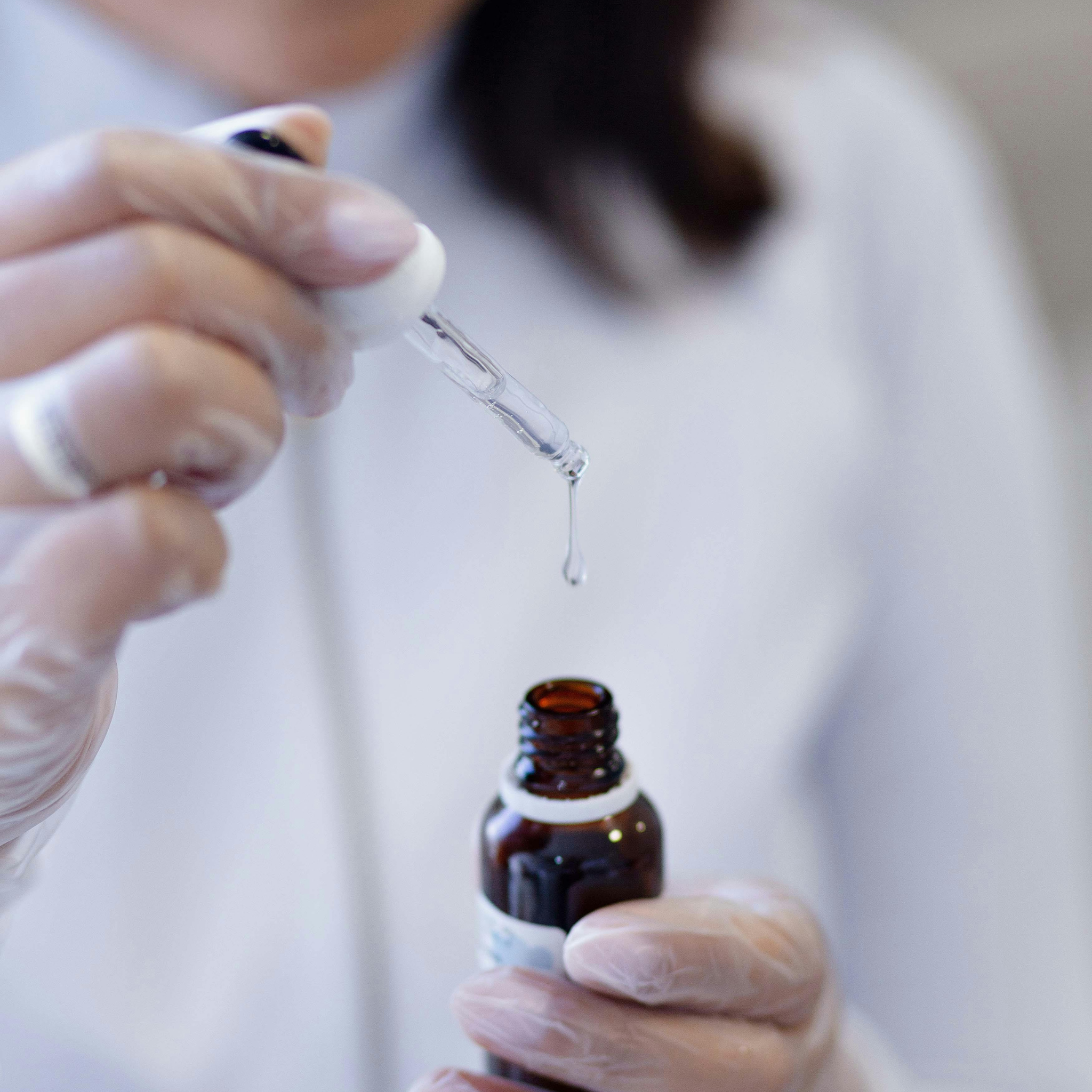 Gloved hands holding a dropper bottle with a drop of liquid.