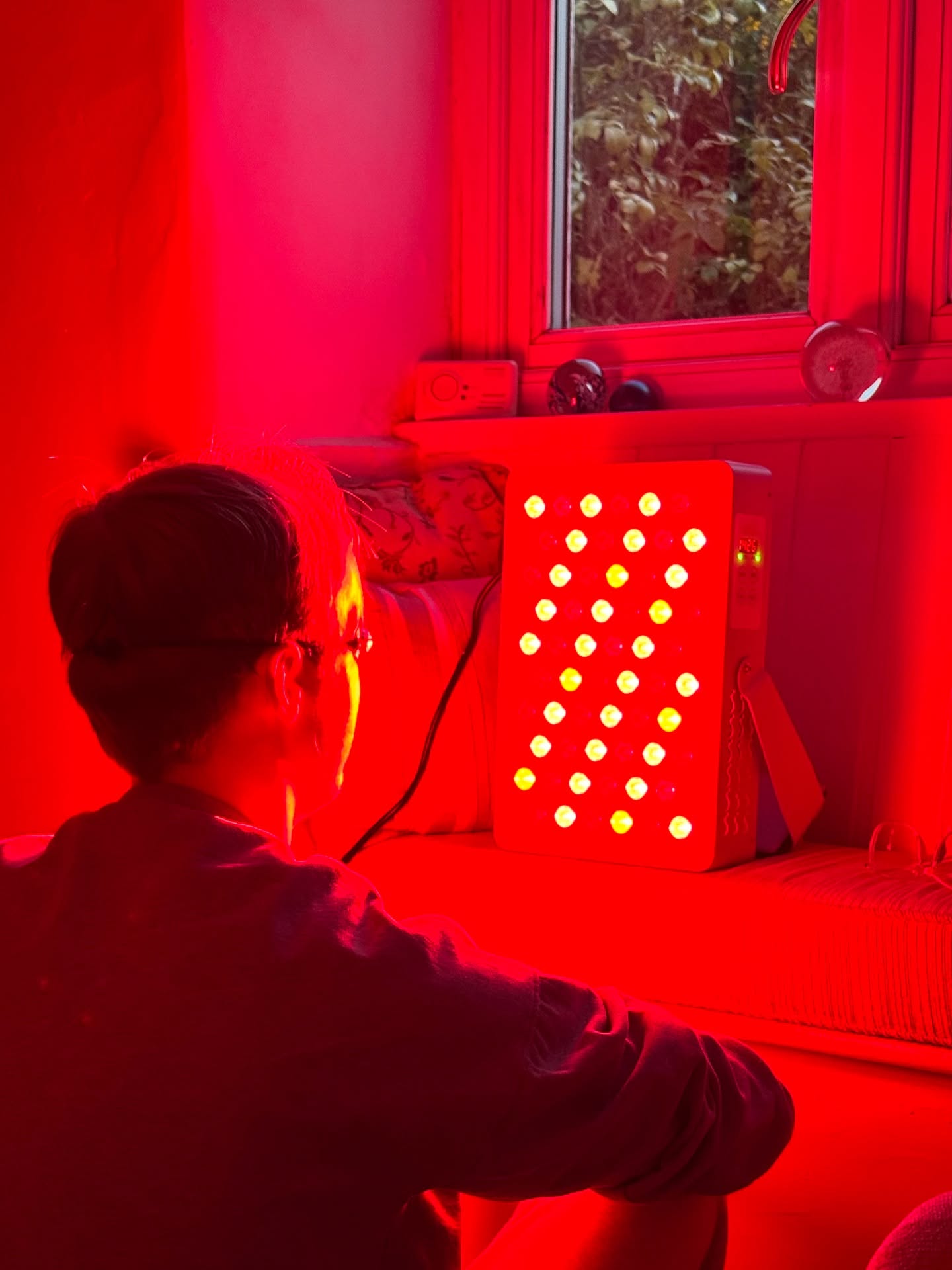 A person, seen from behind, sits in a room facing a red light therapy panel.