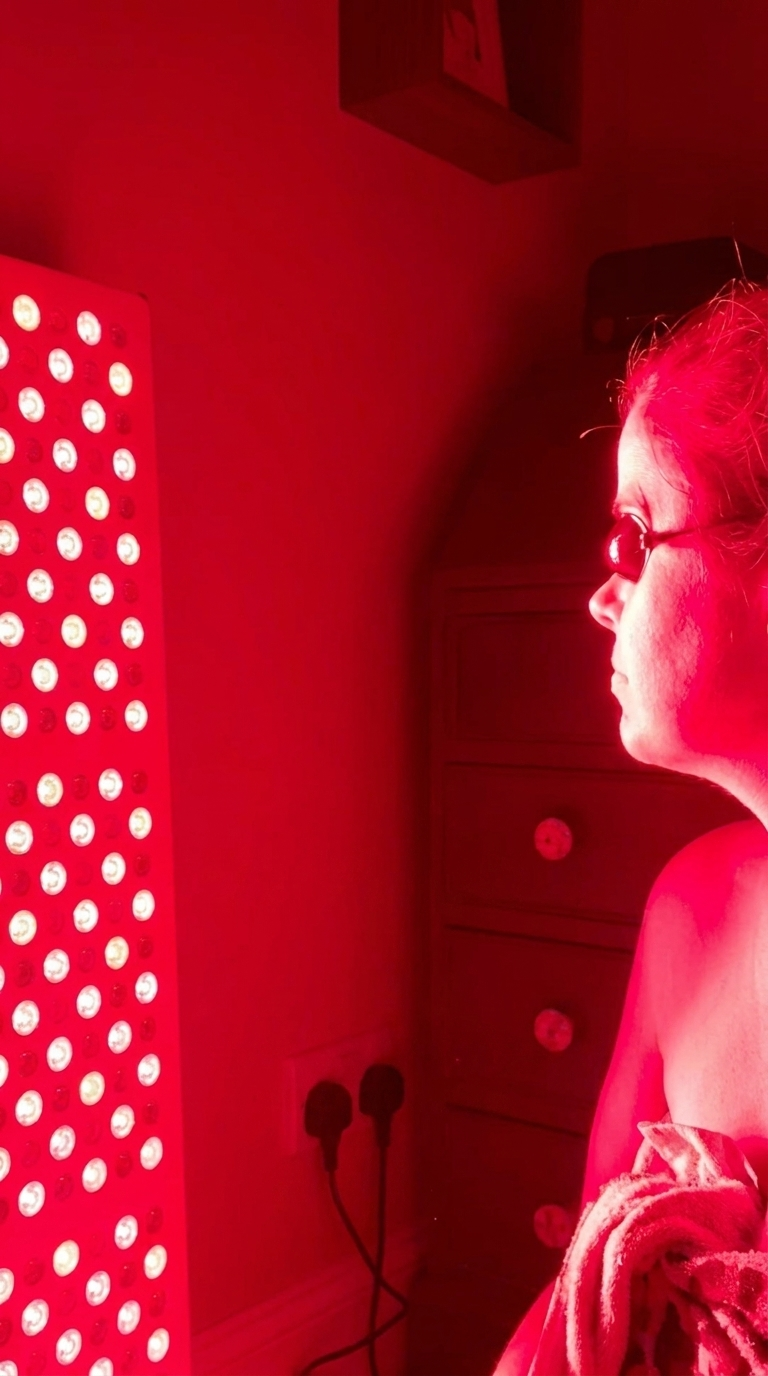 A woman wearing protective eyewear sits in front of a red light therapy panel in a dark room.
