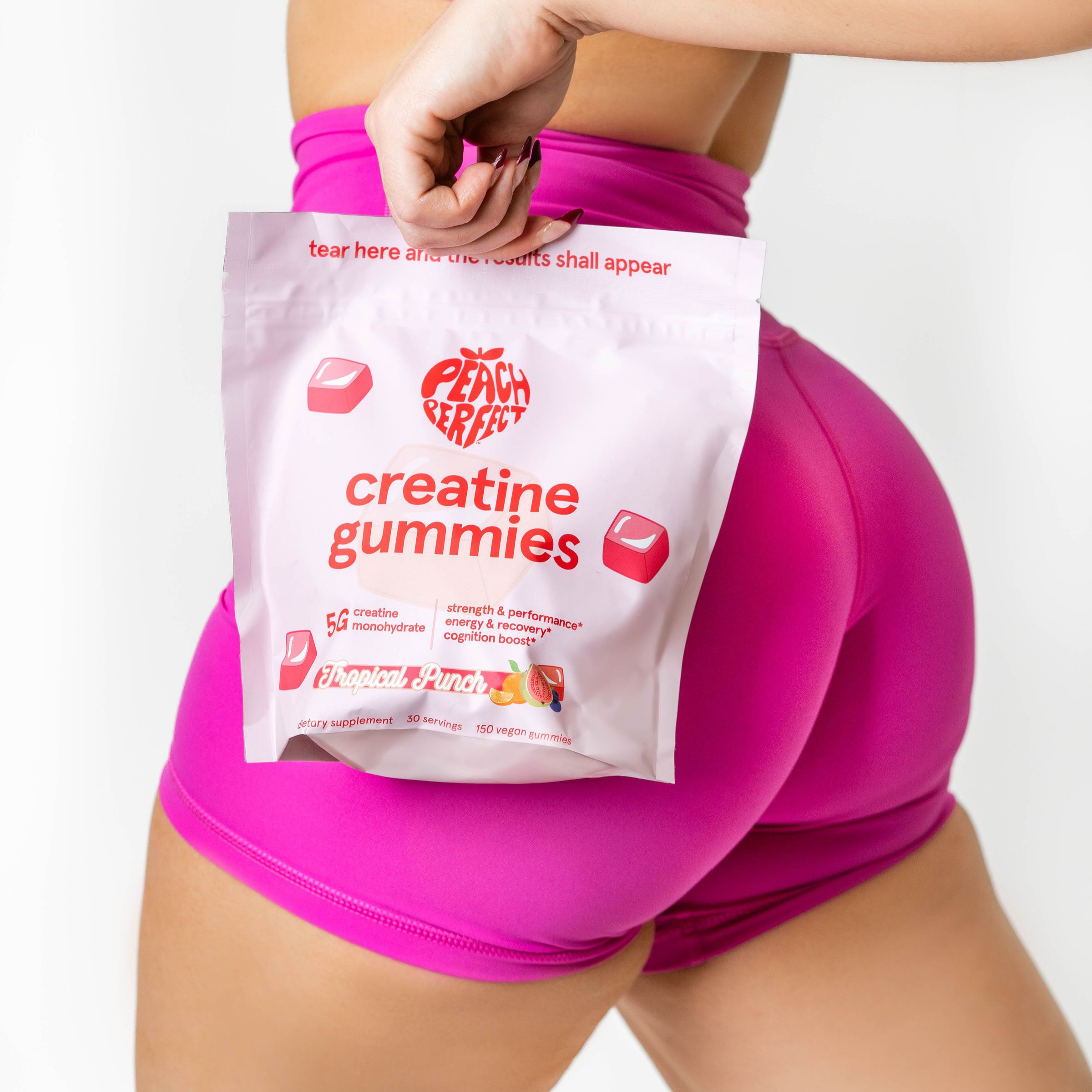 A woman in pink shorts holds a bag of Peach Perfect creatine gummies against her hip.