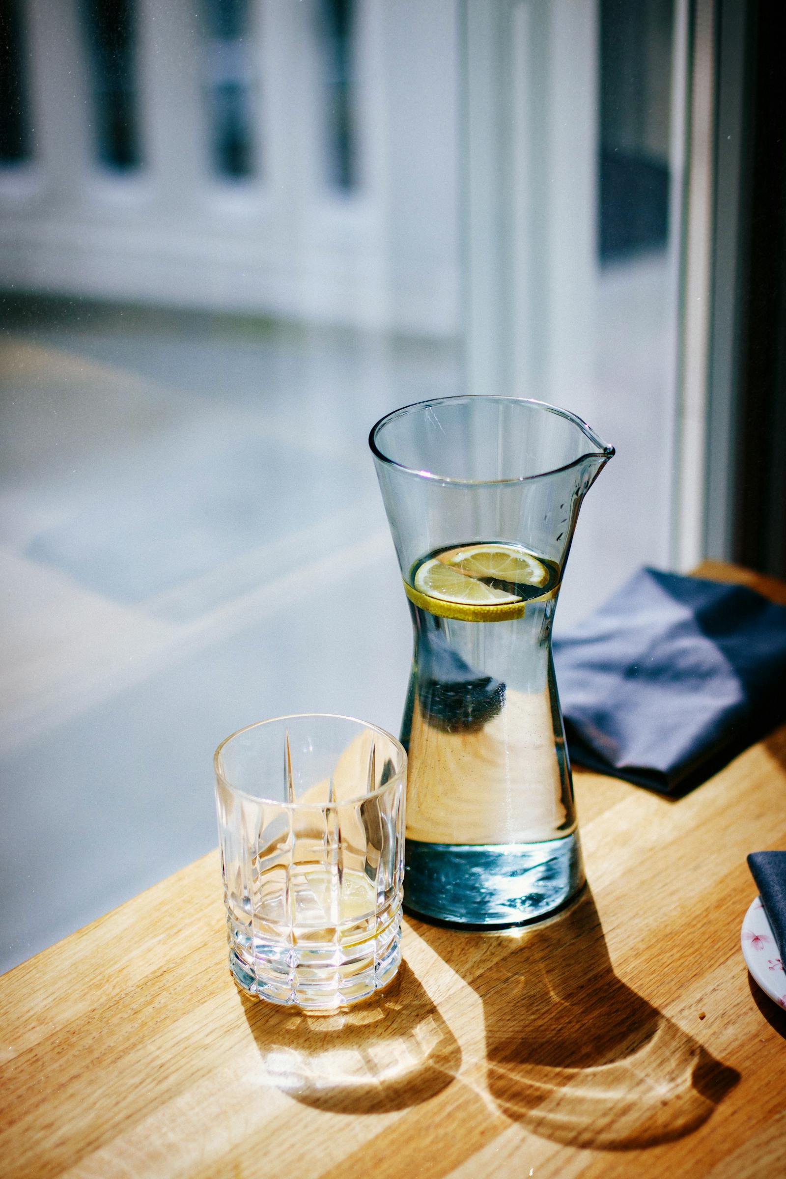 A glass carafe of lemon water and an empty drinking glass sit on a wooden table by a window.