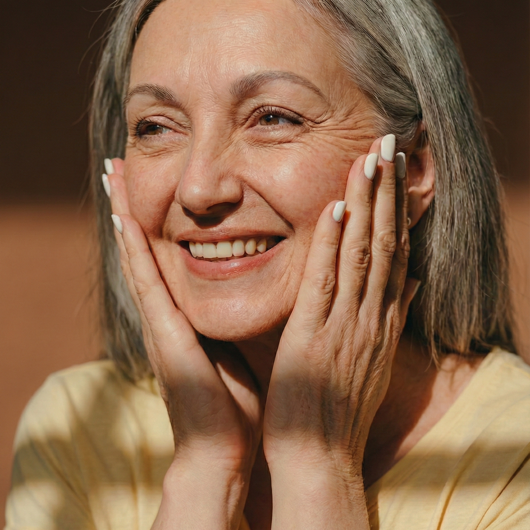 A smiling older woman with gray hair gently holding her cheeks and looking to the side.