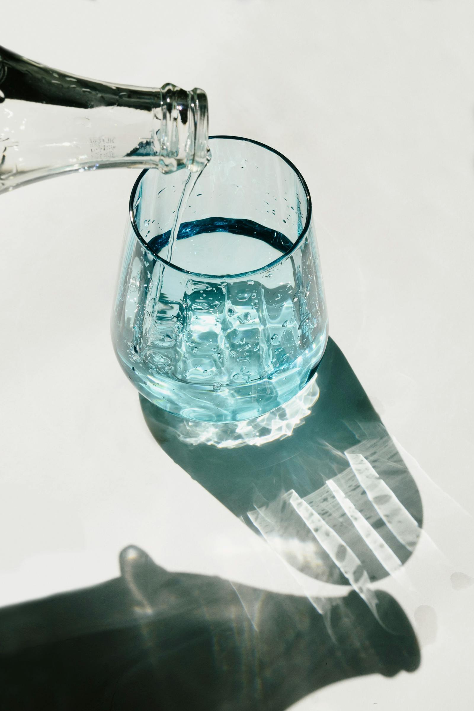 A clear bottle pouring water into a light blue glass tumbler in bright sunlight.