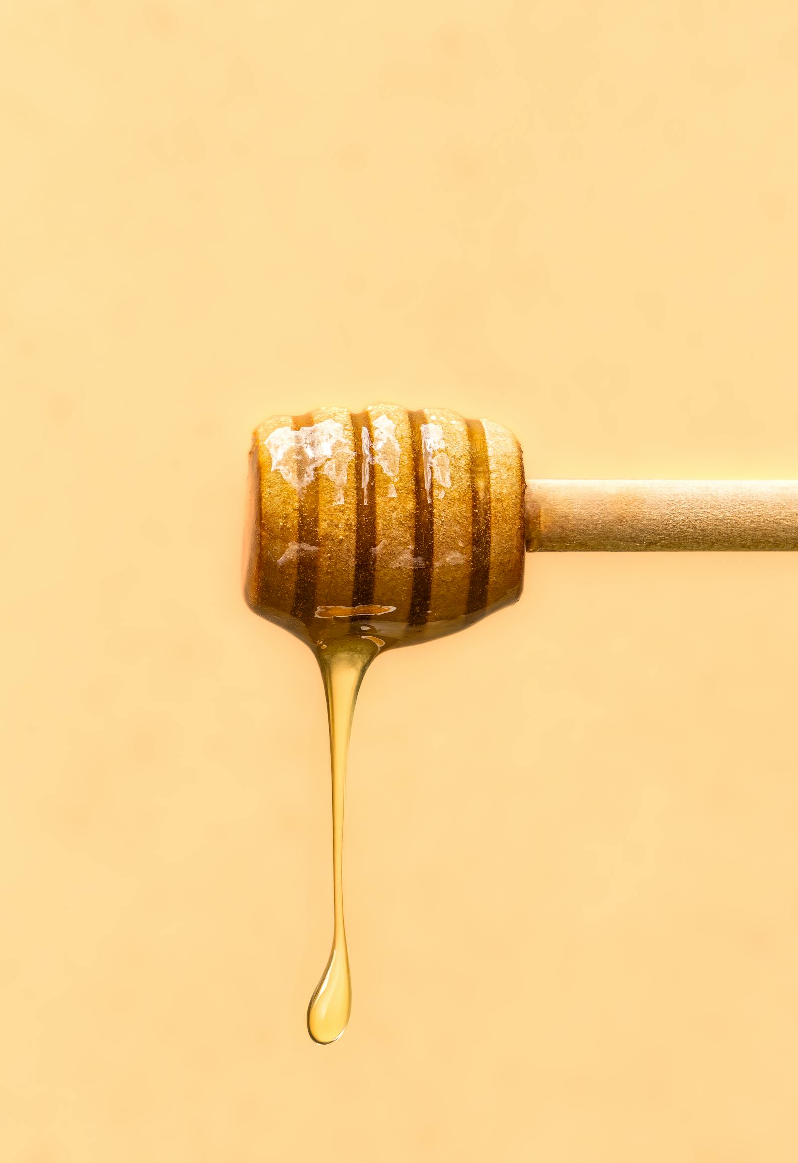 A close-up of a wooden honey dipper with honey dripping from the end against a yellow background.