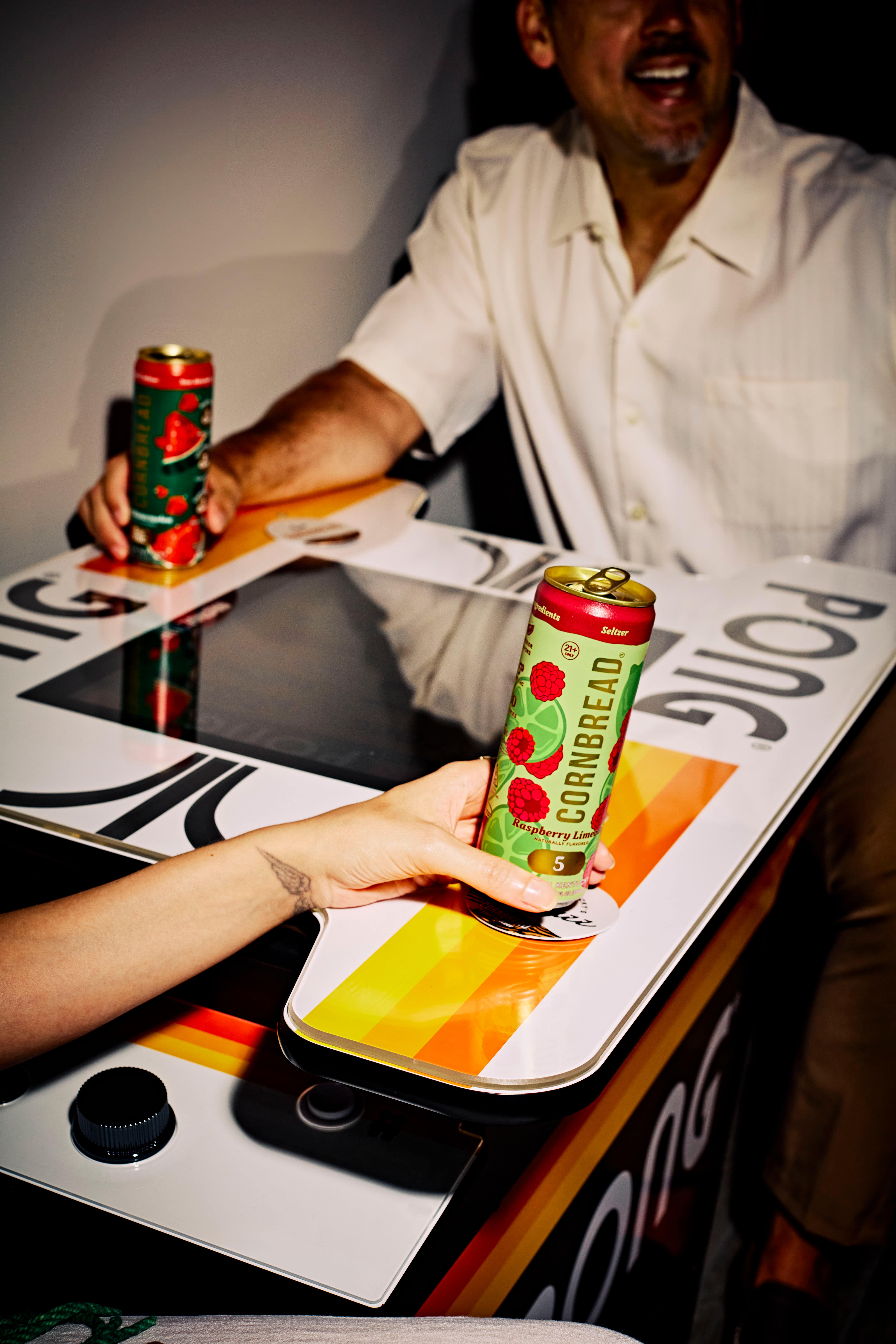 People holding colorful drink cans over a retro tabletop arcade game.