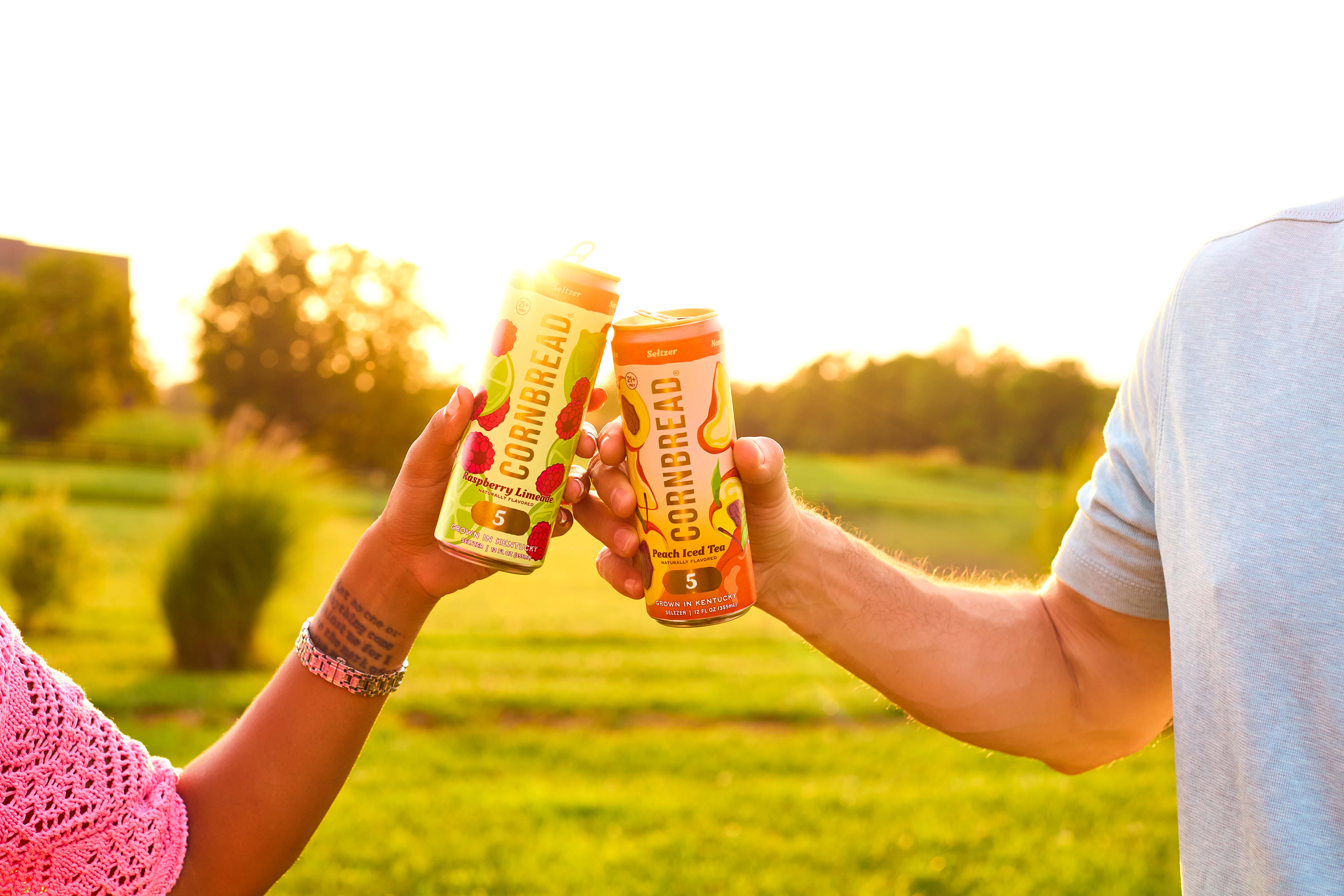Two people toasting with Cornbread seltzer cans in a sunny park.