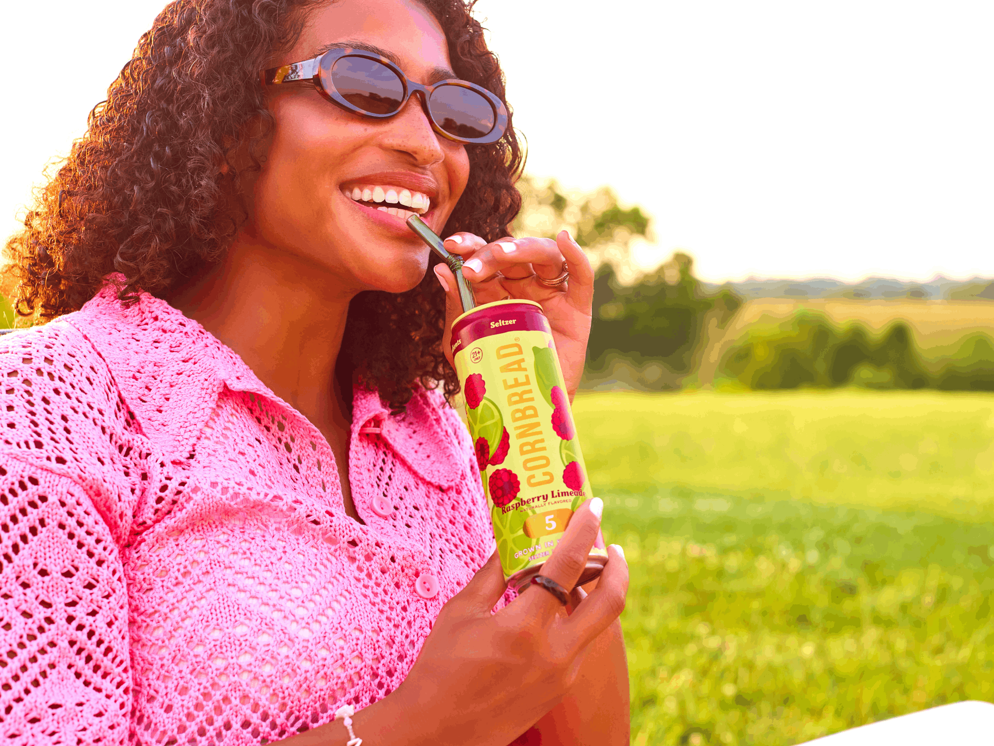 Person in pink sweater drinking from canned beverage with straw outdoors.