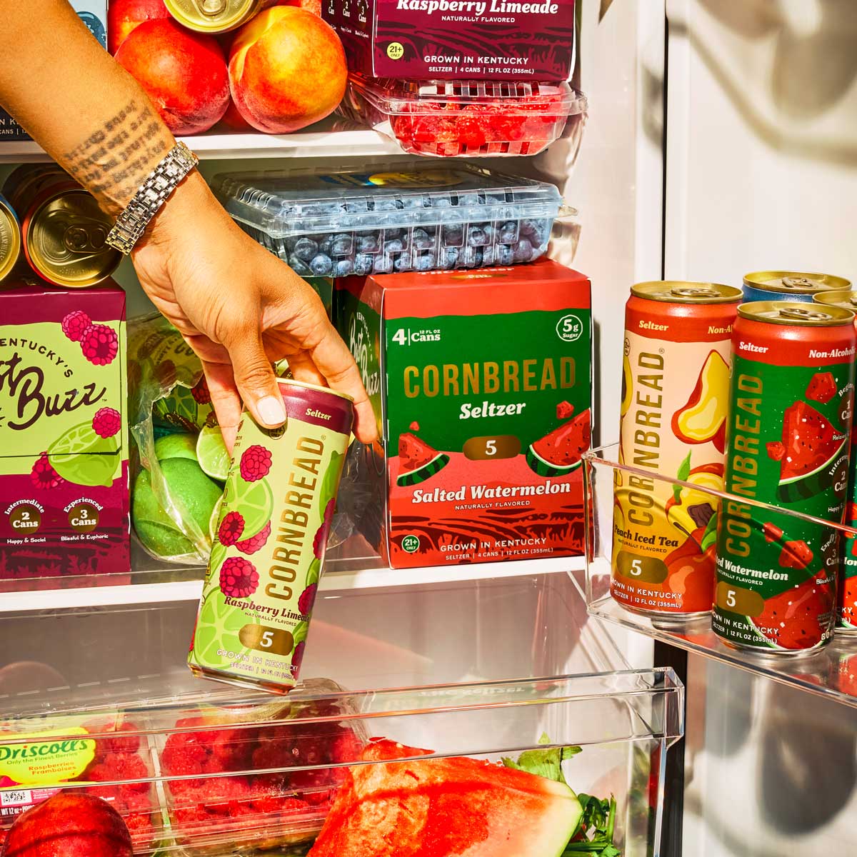 A hand holding a can of cornbread-flavored seltzer in a fruit-filled refrigerator.