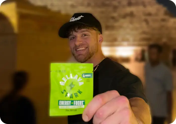 A smiling man in a baseball cap holds up a green packet of Hellava Spark energy supplement.