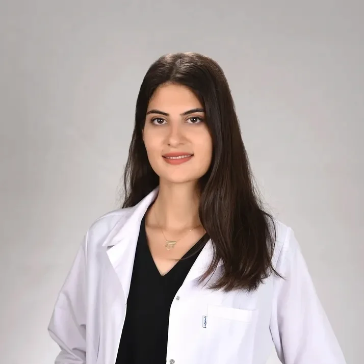 A woman with long dark hair, wearing a white lab coat over a black shirt, smiles at the camera.
