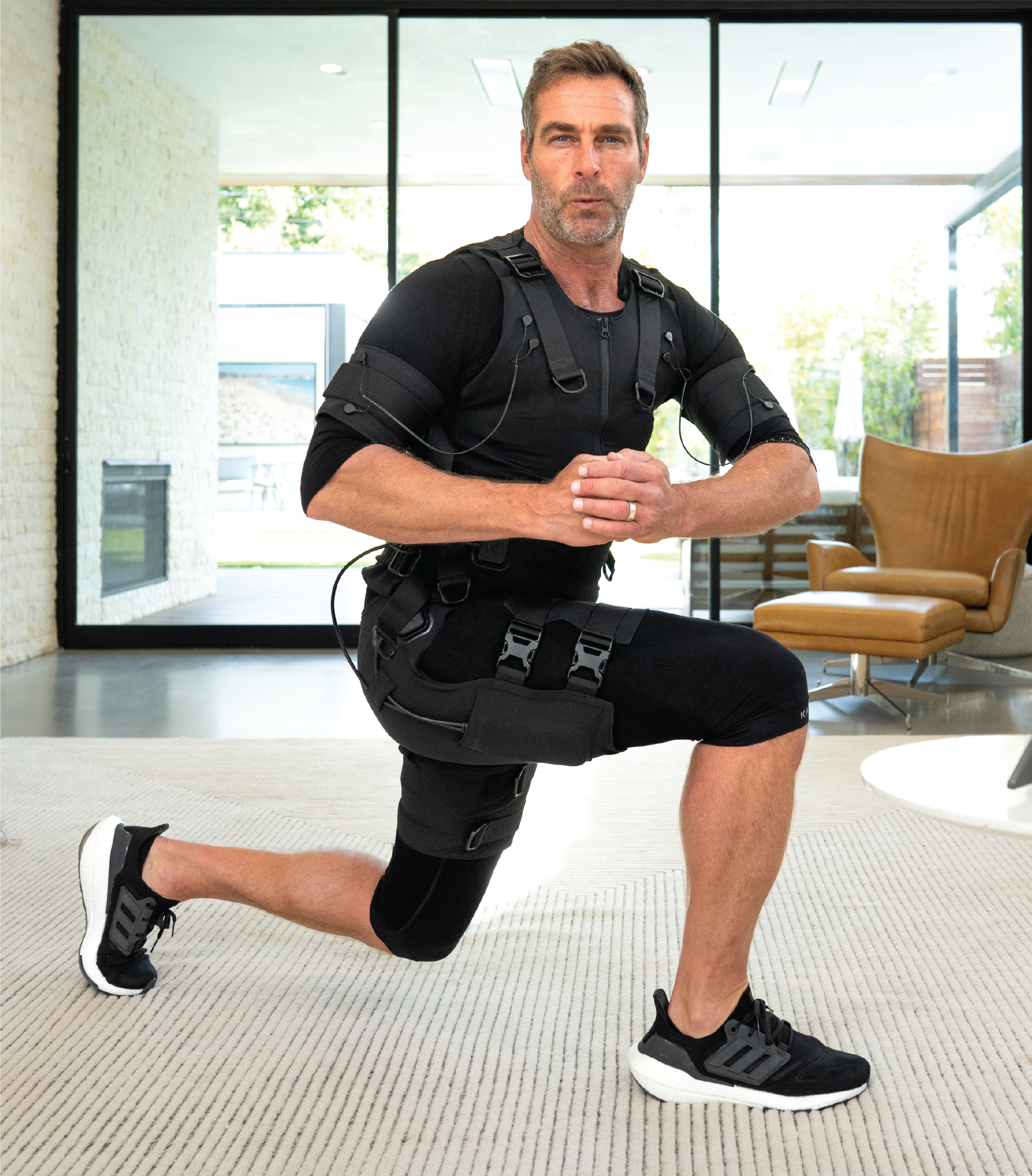 Man in exercise suit kneeling indoors, wearing black sneakers.