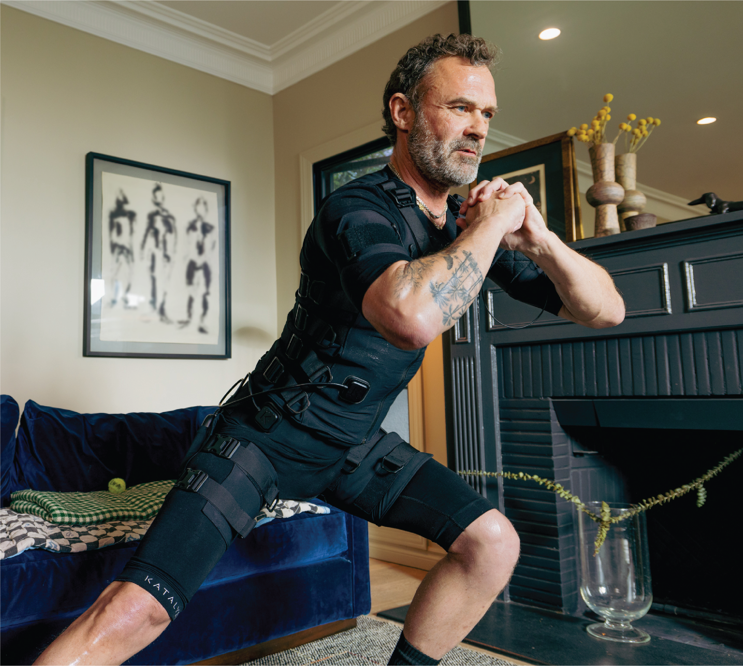 Man exercising in a living room wearing a black suit with straps.