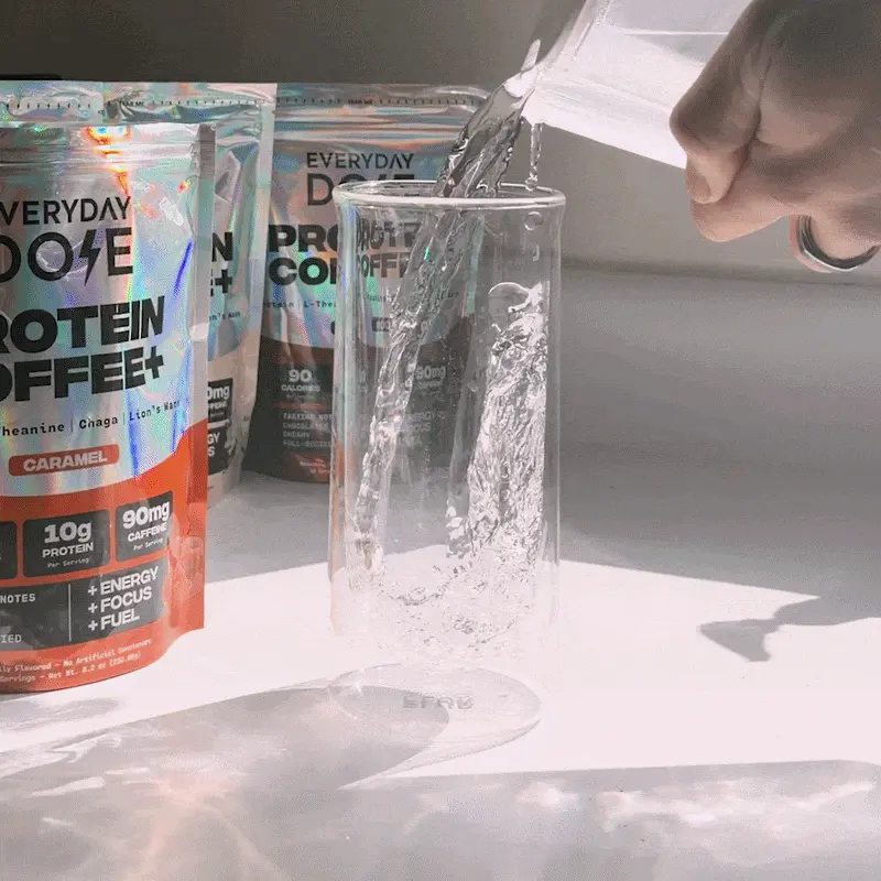 A hand pours water into a tall glass, with bags of Everyday Dose Protein Coffee+ in the background.