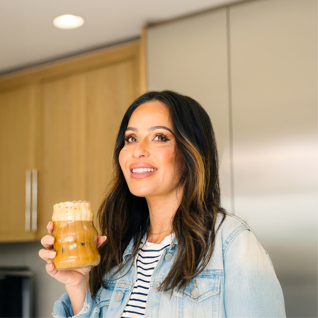 Person smiling, holding a glass of iced coffee in a kitchen.