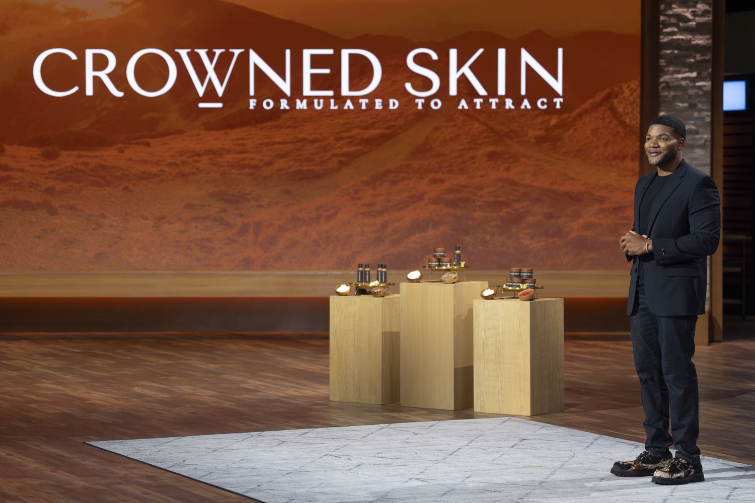 A man stands on a stage next to pedestals displaying Crowned Skin products.