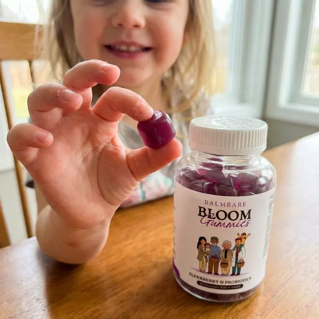 A young girl holds up a purple gummy with a bottle of Balmbare Bloom Gummies on the table.