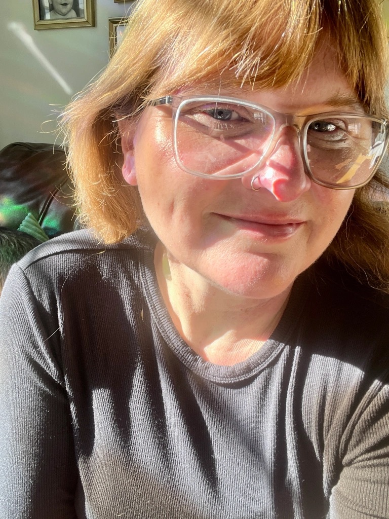 Person with glasses smiling in sunlight, wearing a dark shirt.
