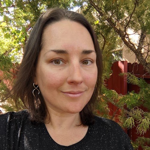 Person with dark hair smiling outdoors near trees and a red fence.