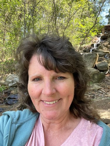 Woman smiling outdoors with trees and rocks in the background.