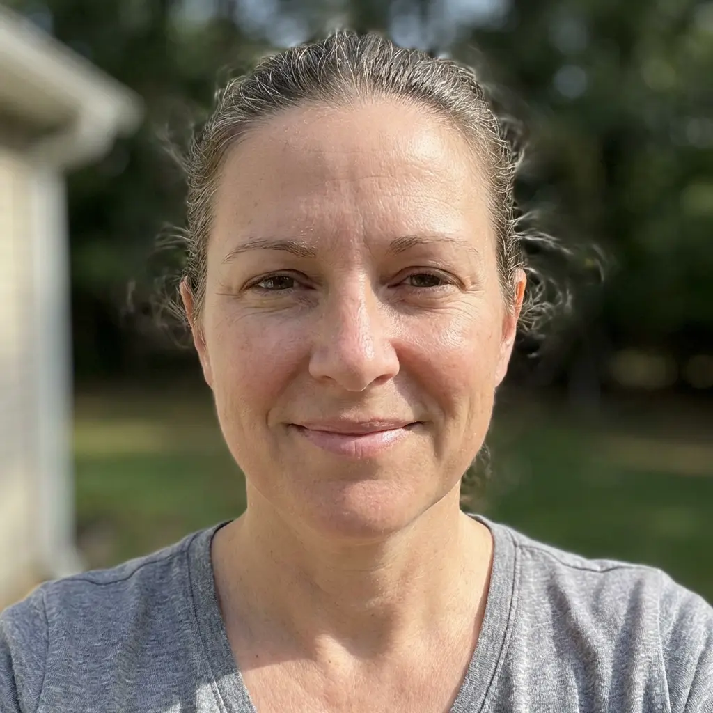 A headshot of a woman with brown hair pulled back, wearing a grey t-shirt and smiling outdoors.