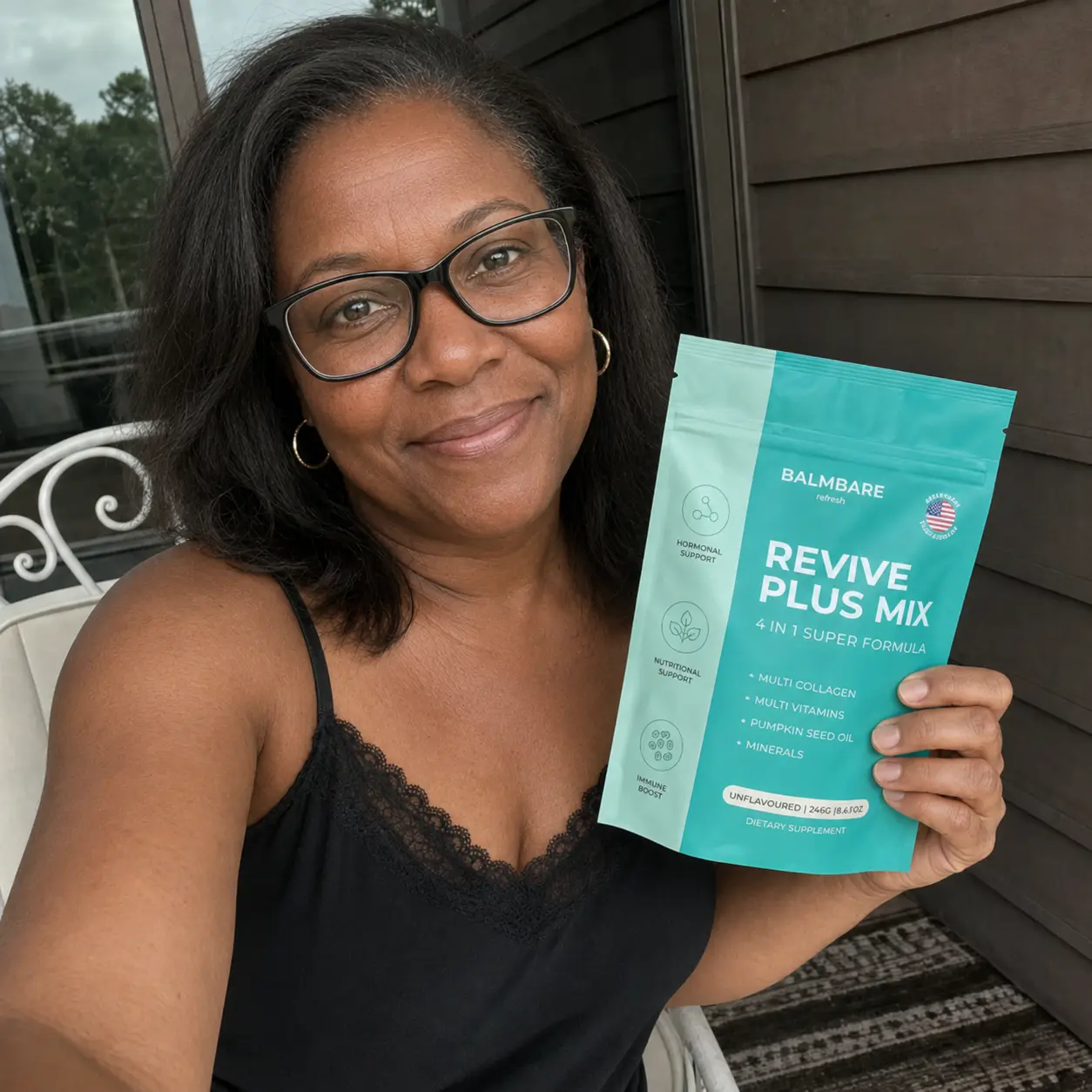 A smiling woman with glasses, wearing a black top, holds a pouch of Balmbare Revive Plus Mix.