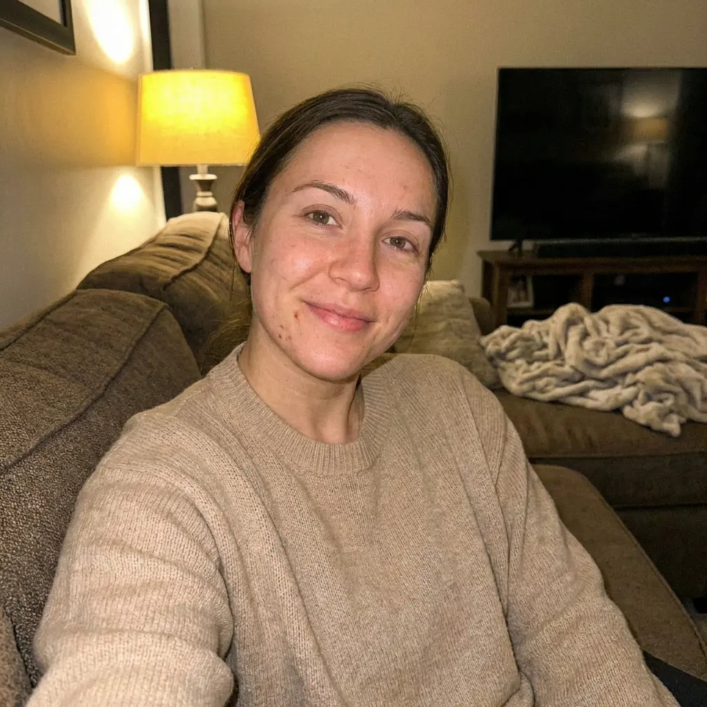 A woman in a tan sweater sits on a brown couch in a dimly lit room and smiles.