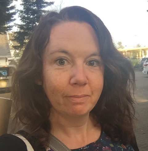 A woman with curly hair standing outdoors, with trees and cars in the background.