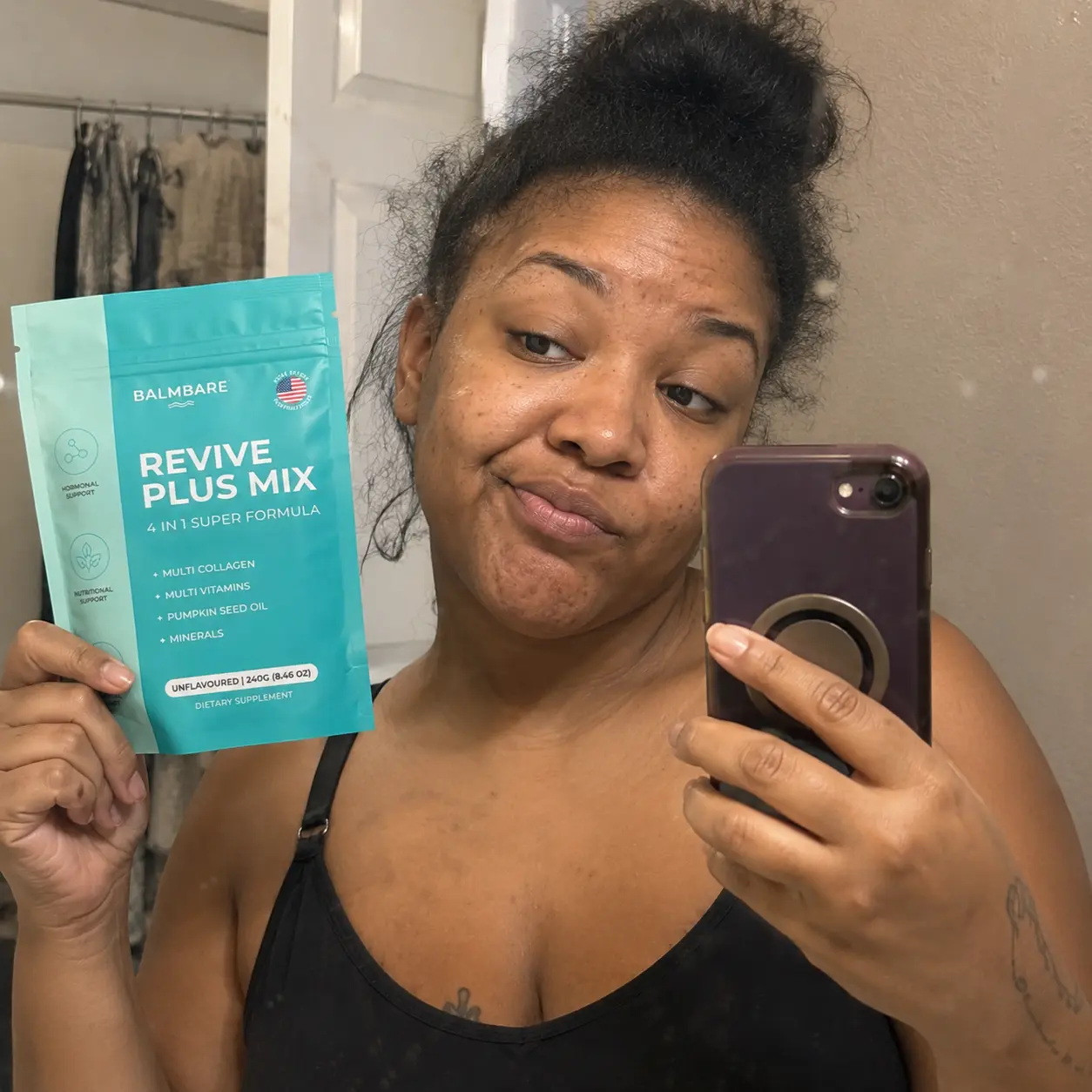 A woman takes a selfie in a mirror while holding up a pouch of Balmbare Revive Plus Mix.