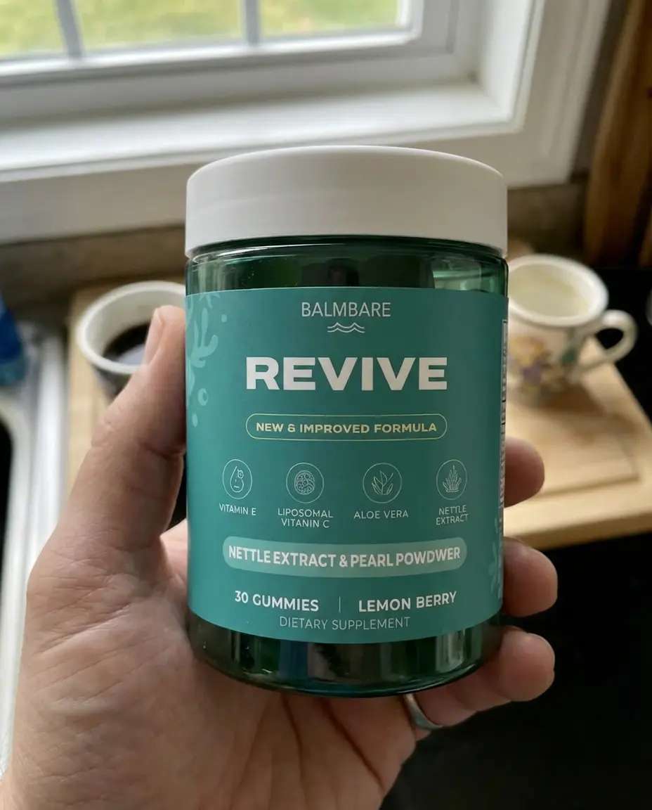 A hand holding a green jar of Balmbare Revive dietary supplement gummies in a kitchen setting.