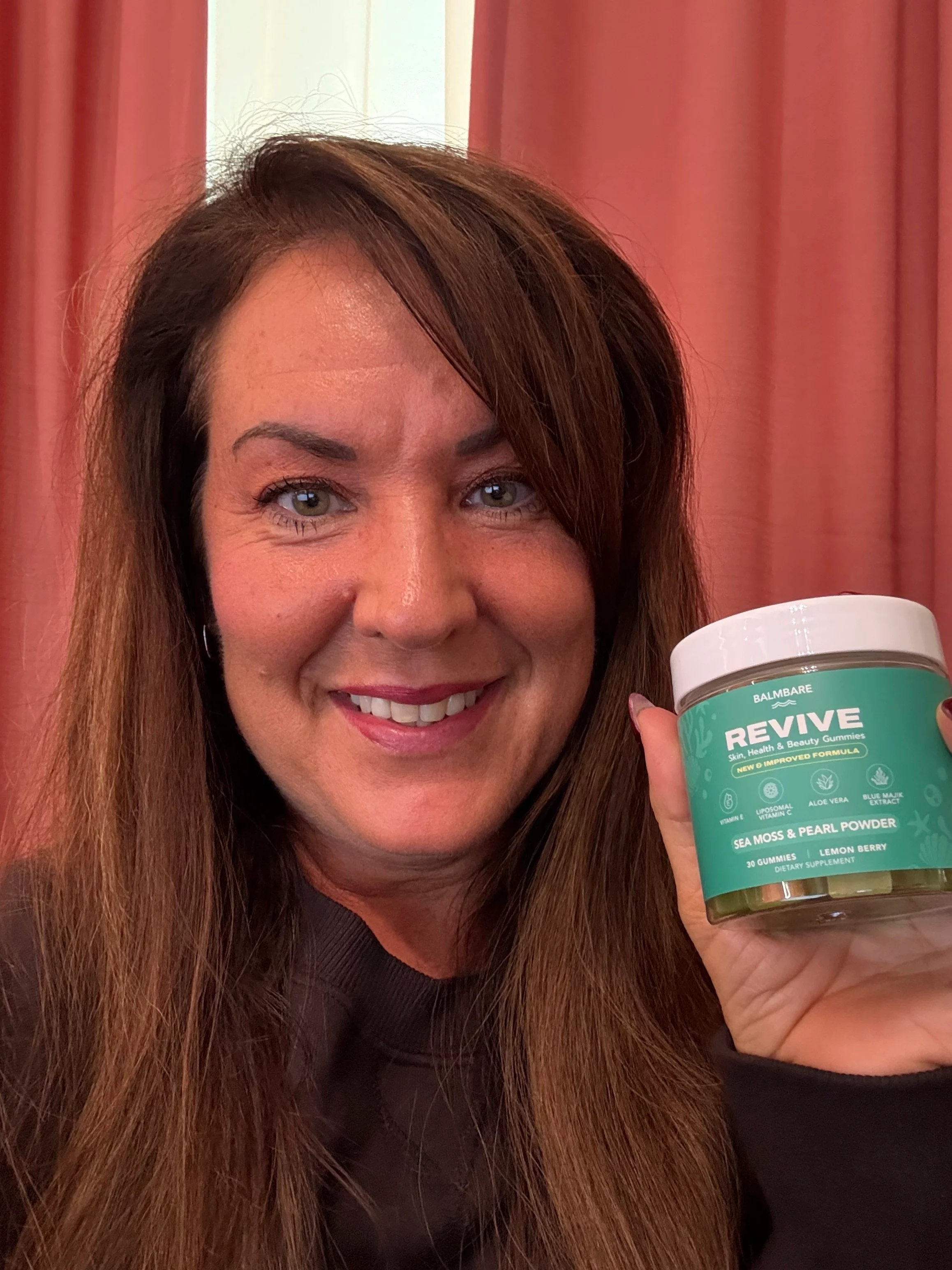 Person holding a jar of 'Revive' gummies, smiling in front of pink curtains.