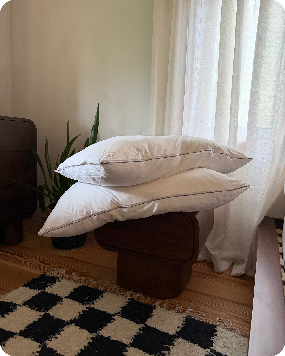 Two white pillows stacked on a wooden nightstand in a bedroom with a checkered rug.