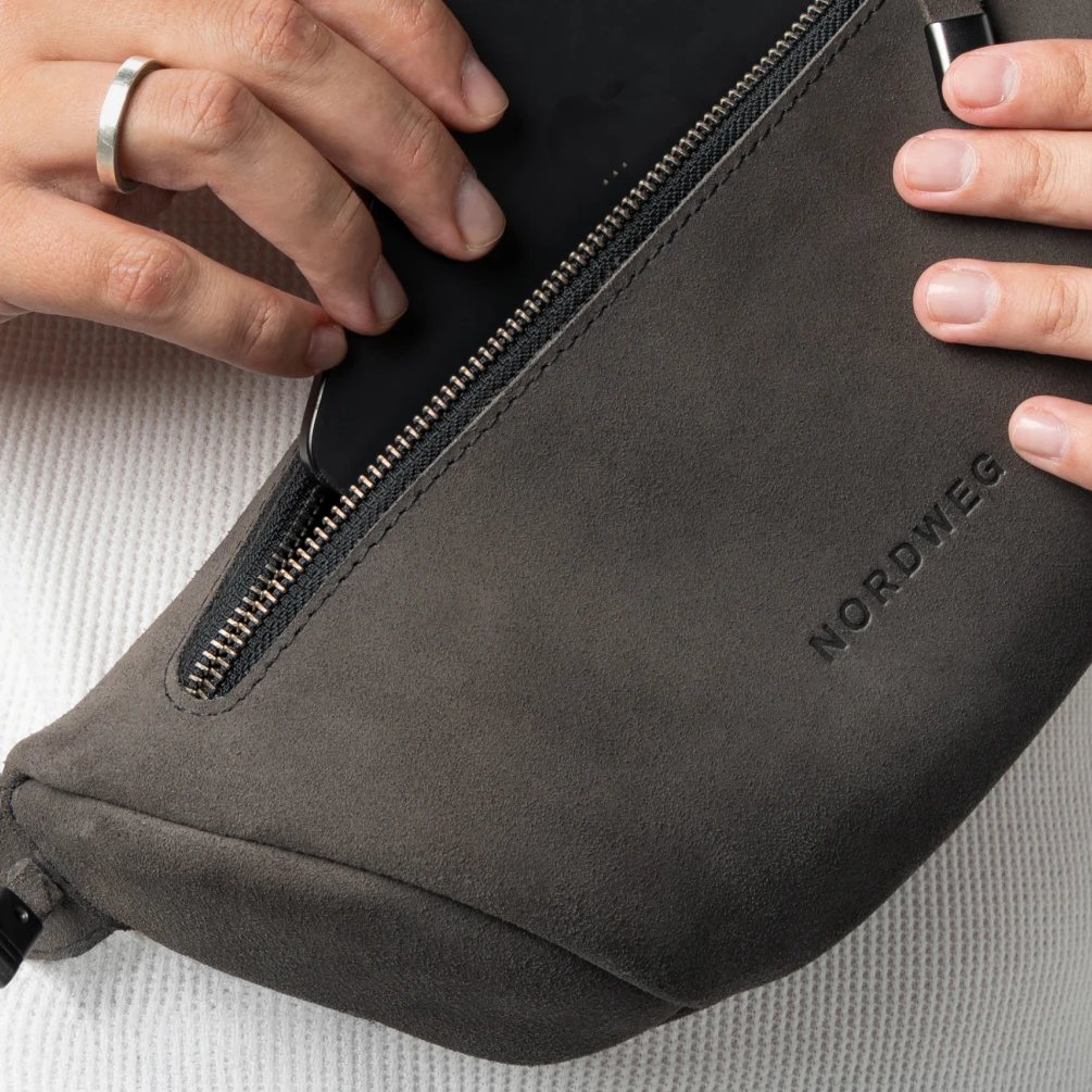 Mãos fecham o zíper da Sling Bag Pulse Limited Edition Nordweg em couro camurça na cor Dark Steel