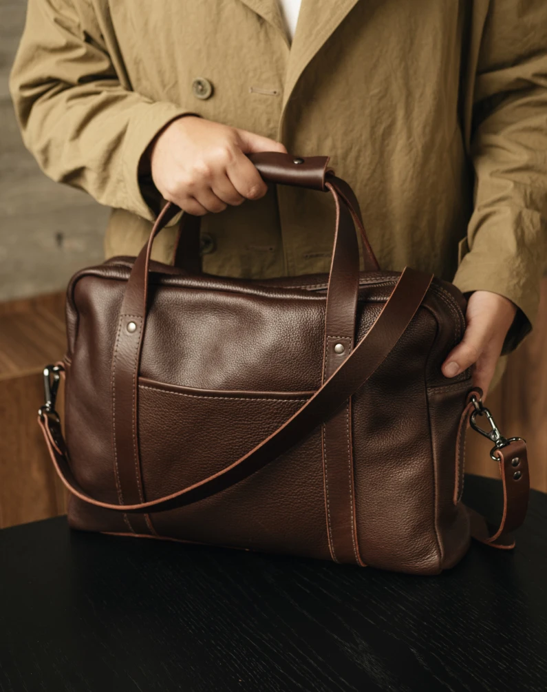 A person in a tan jacket holds a brown leather bag by its handle on a black table.