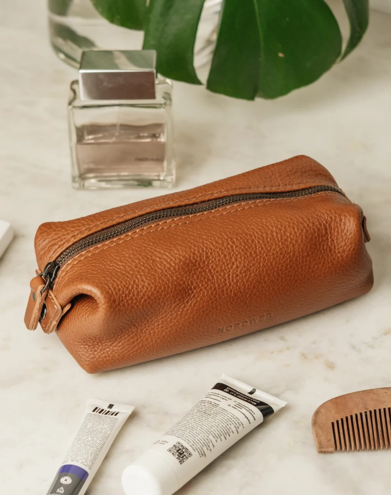 A brown leather toiletry bag on a marble surface with grooming products like cologne, lotions, and a comb.