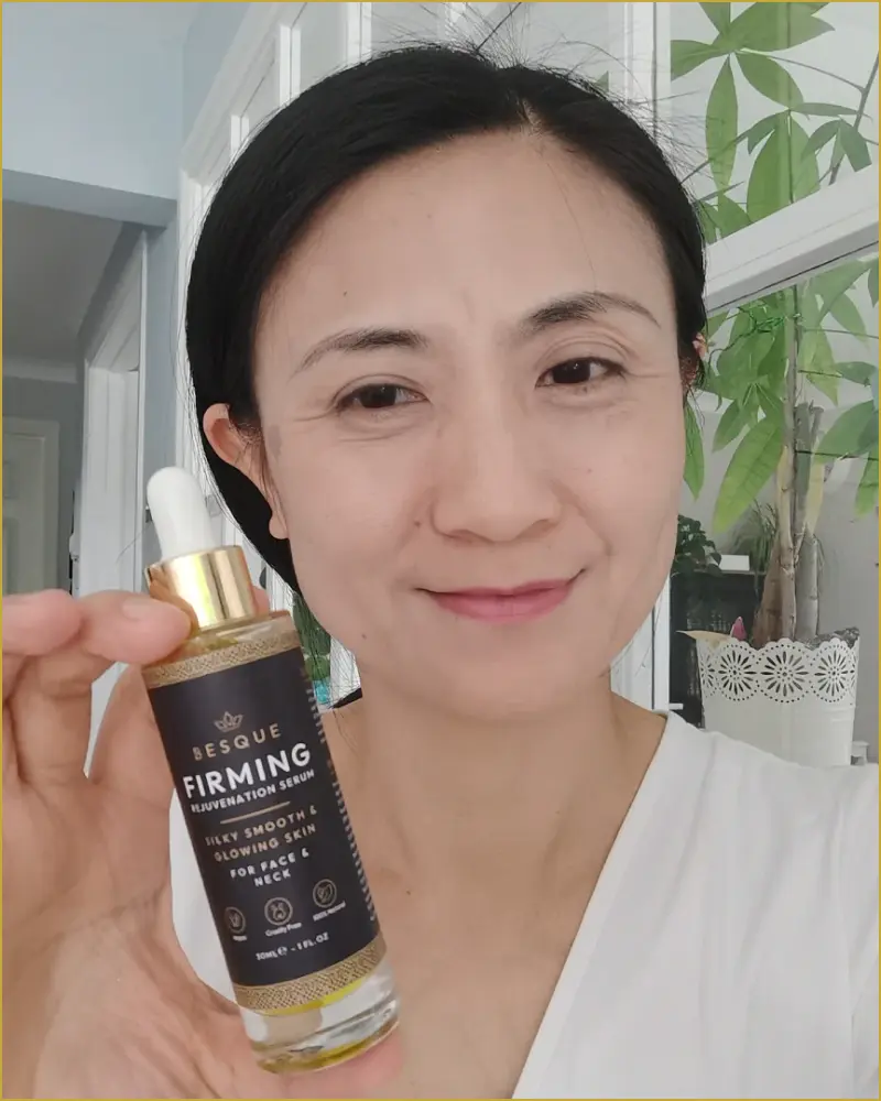Person holding a bottle of firming rejuvenation serum, standing indoors with plants visible in the background.