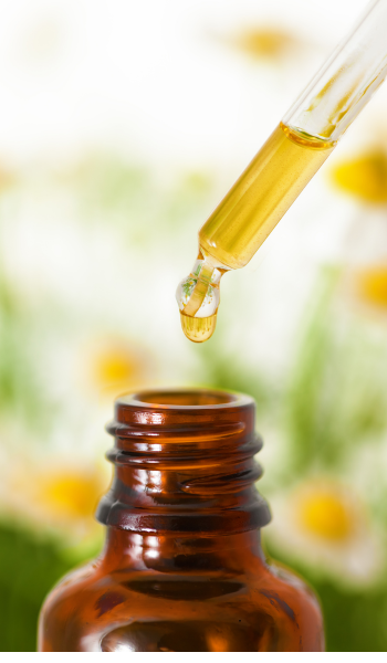 Dropper with liquid above a brown bottle, blurred flowers in background.