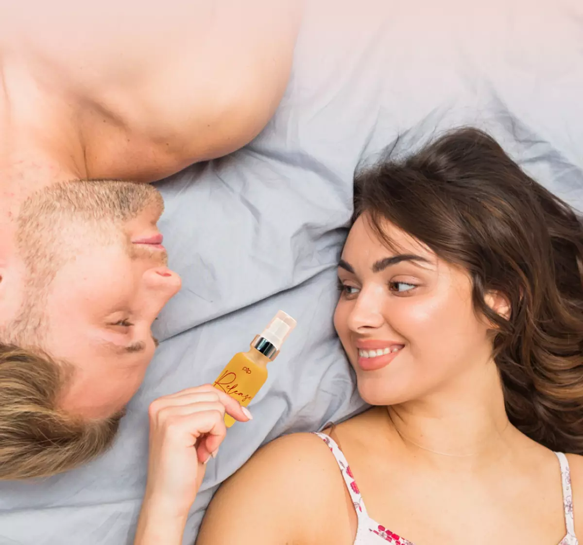 A man and a woman lie in bed, looking at each other, as she holds up a small bottle.