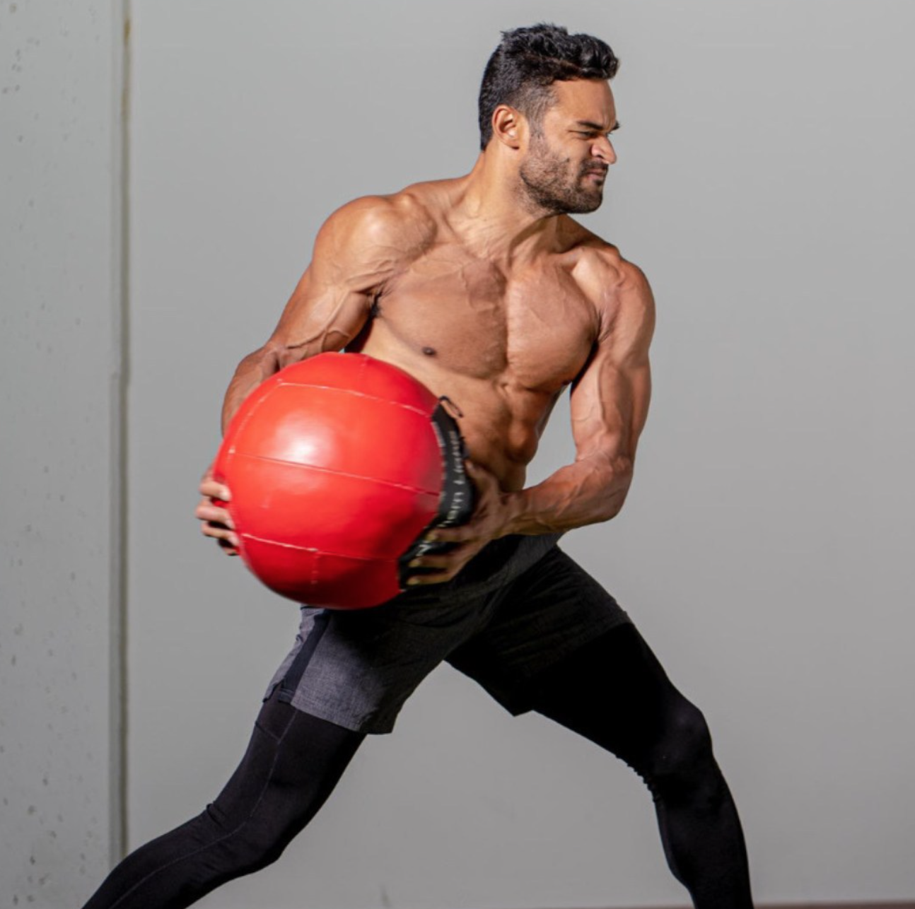 A muscular, shirtless man twists his torso while holding a large red exercise ball against a grey background.